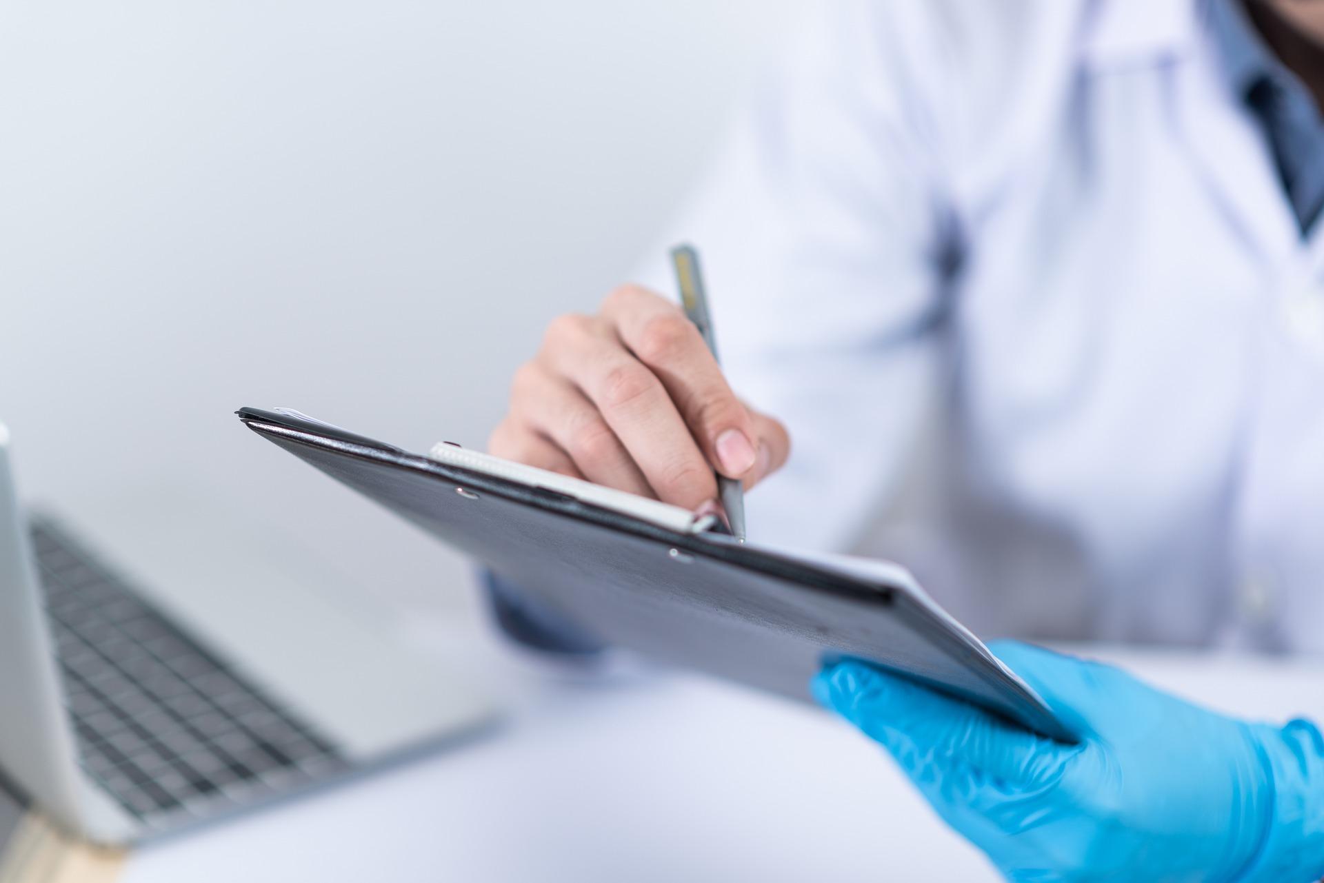 Healthcare worker in blue gloves writing on a clipboard next to a laptop