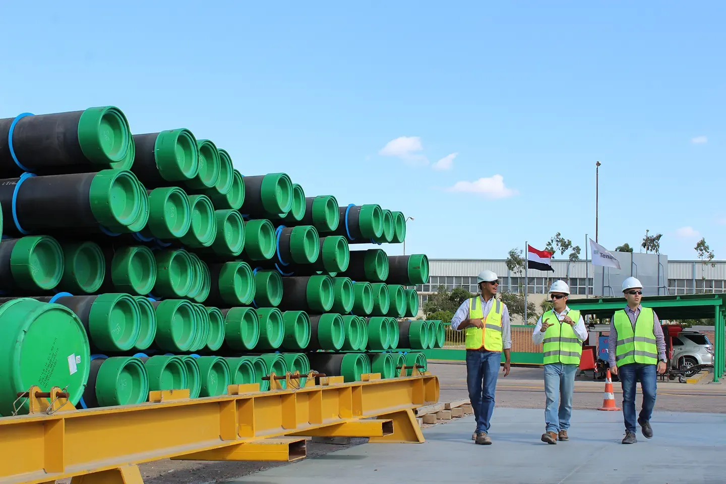 Engineers in hard hats walking past stacked capped steel pipes at a Tenaris plant